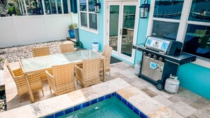 A patio with a glass-top table and wicker chairs, a small pool with blue tiles, and a barbecue grill.