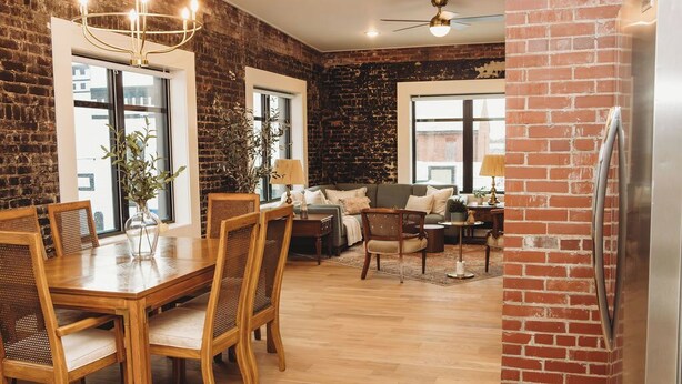Een ruime woonkamer met een bakstenen muur, een eethoek met een houten tafel en stoelen, en een moderne keuken met een koelkast.