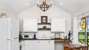 Una cocina con armarios blancos, microondas, lavamanos y comedor con mesa y sillas de madera.