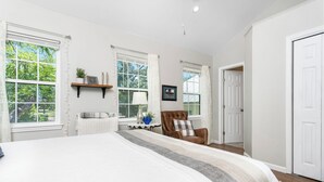 Un dormitorio amplio con una cama grande, dos ventanas con cortinas, una silla, una mesita de noche con lámpara y una puerta.