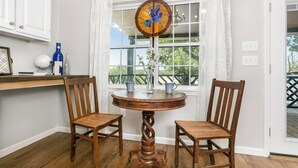 Un comedor con una mesa redonda de madera y dos sillas, una ventana con cortinas blancas y vistas a la vegetación exterior.