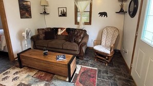 Una sala de estar con un sofá marrón, una mesa de centro de madera, un tapete estampado, una silla y un ventanal con persianas.