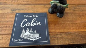Una mesa de madera con un cartel de bienvenida para el libro de visitas de la cabaña y una figurita de oso sosteniendo un pez.