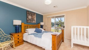 A bedroom with a bed, two bedside tables, a chair, a crib, and a large window. - Palms on Collingwood by Experience Jervis Bay (Vincentia)