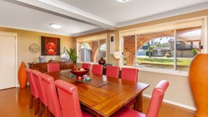 A dining room with a wooden table, red chairs, and a view of a brick building through large windows. - Palms on Collingwood by Experience Jervis Bay (Vincentia)