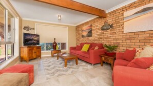 A living room with a red sofa, a wooden coffee table, a TV on a wooden cabinet, and a brick wall with artwork. - Palms on Collingwood by Experience Jervis Bay (Vincentia)
