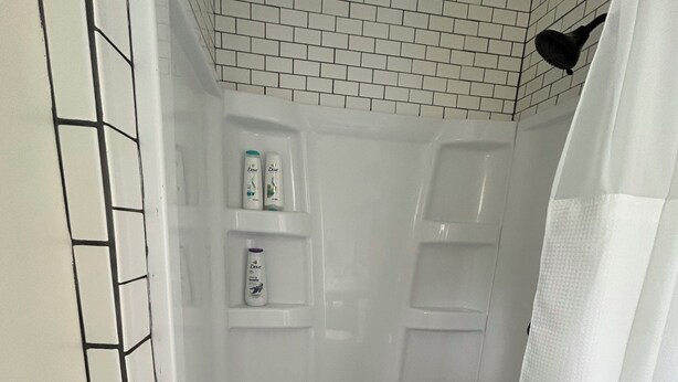 A small, white-tiled shower stall with a shower curtain, a shelf with toiletries, and a drain in the center. - Minutes from Marquette, close to Munising, great location for exploring the UP! (Skandia)