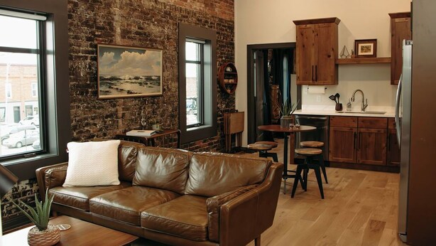 A cozy living room with a leather sofa, a coffee table, and a brick wall. There is a kitchen area with wooden cabinets and a refrigerator. - The Aaron is a charming 2-bedroom/ 2 bathroom apartment.  (Leitchfield)