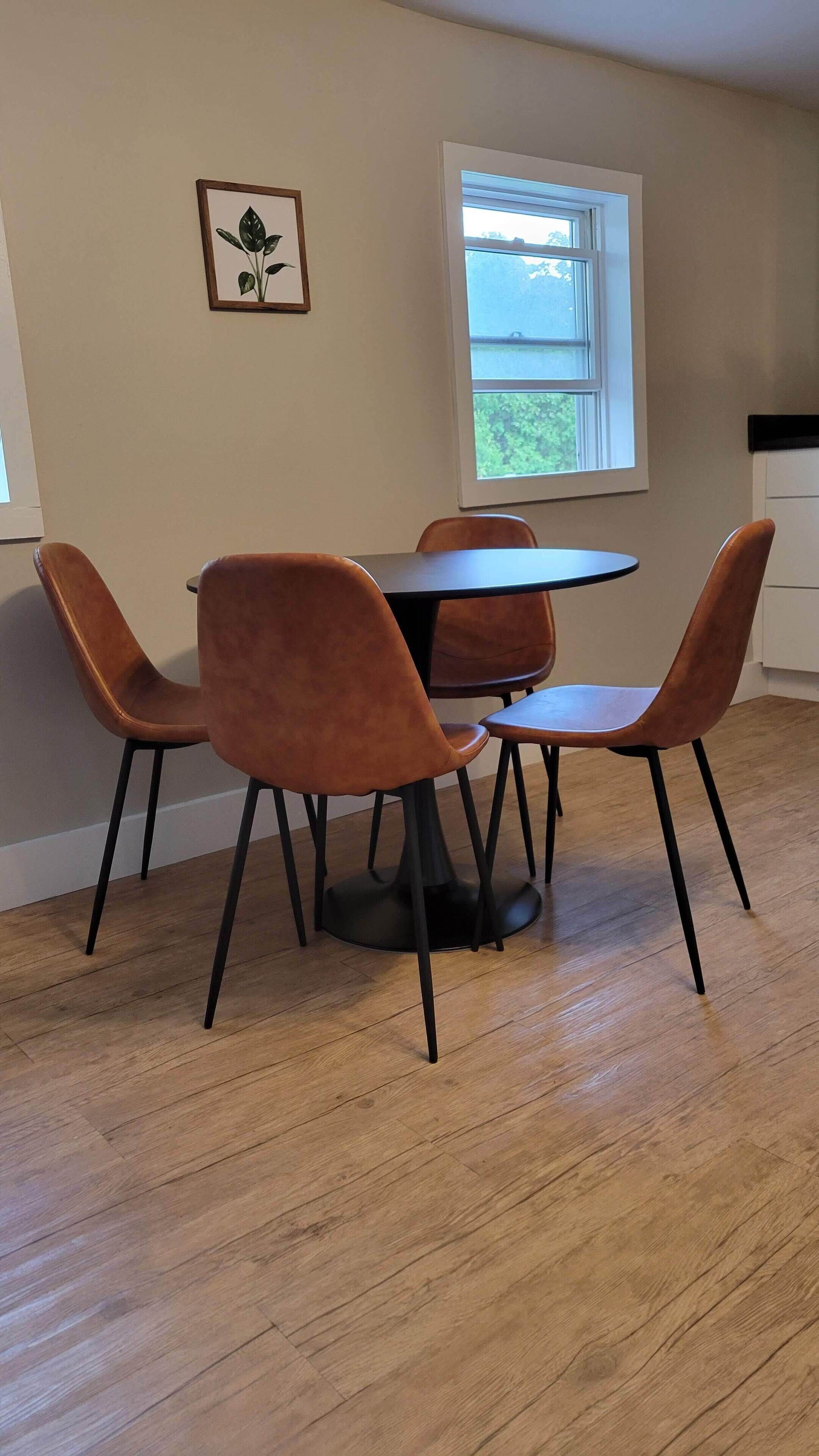 Un comedor con una mesa redonda y cuatro sillas, suelo de madera, una ventana y un cuadro en la pared.