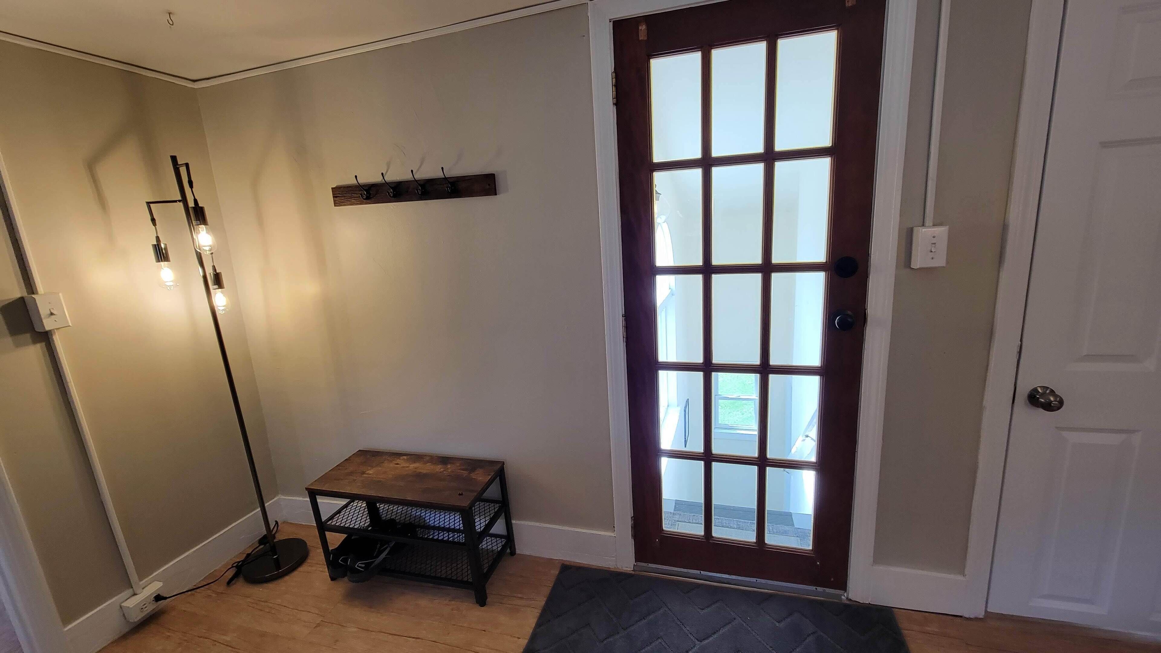 A hallway with a wooden floor, a dark brown door with glass panels, a small table with a metal frame, and a black rug at the door's entrance.
