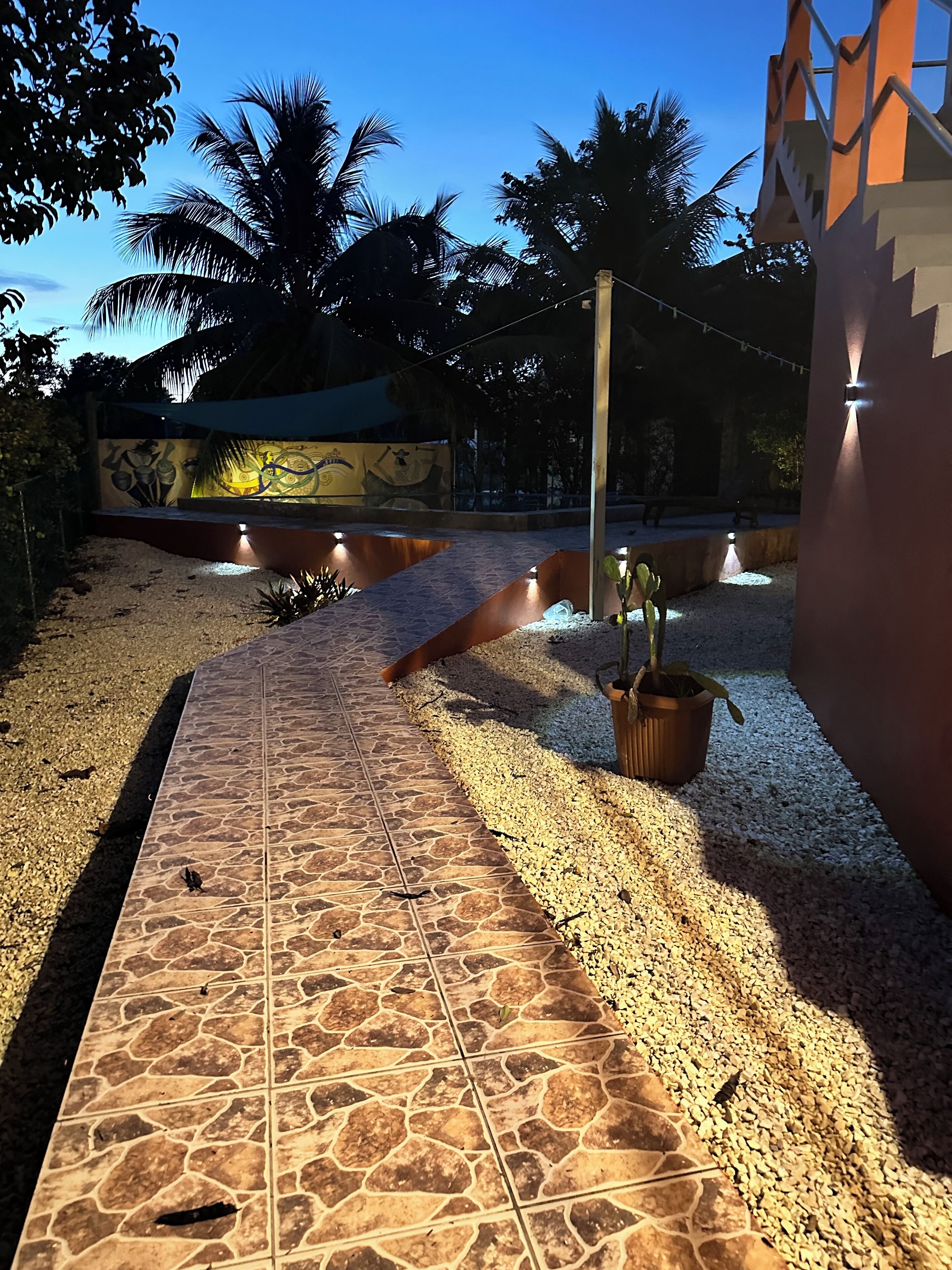 A pathway with stone tiles leading to a seating area with a mural, surrounded by palm trees and potted plants.