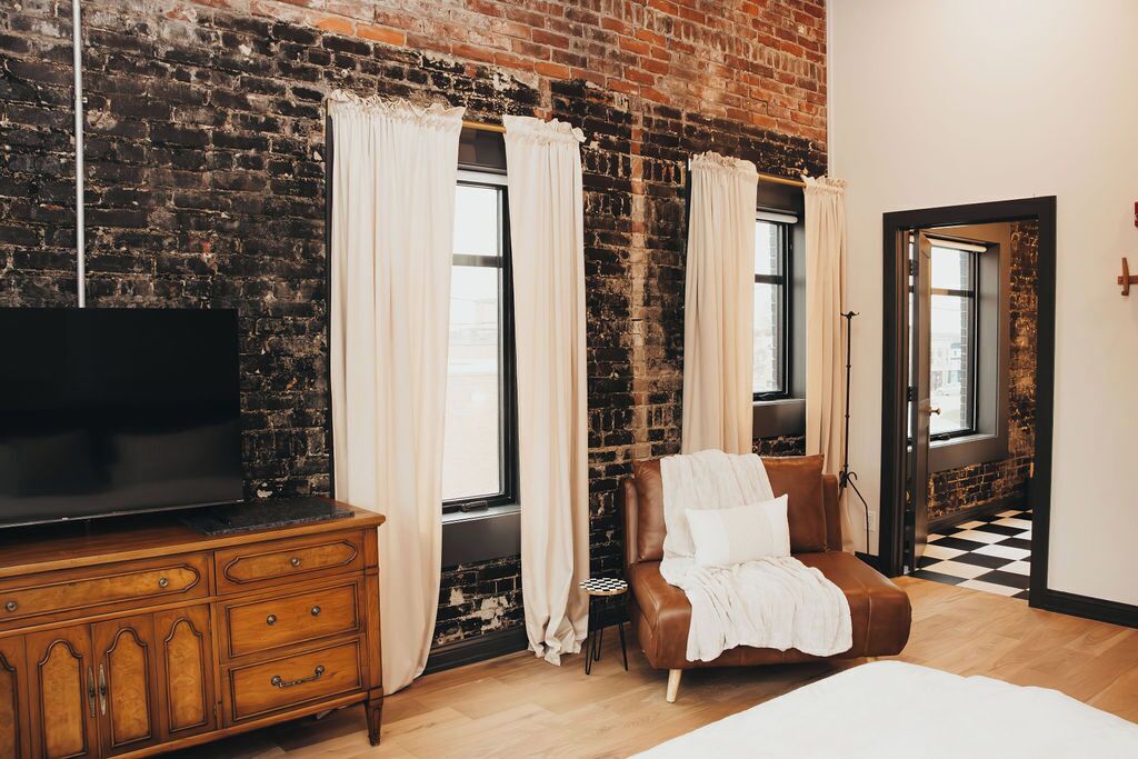 A room with a brick wall, a wooden dresser with a TV, a brown leather chair, and a bed.