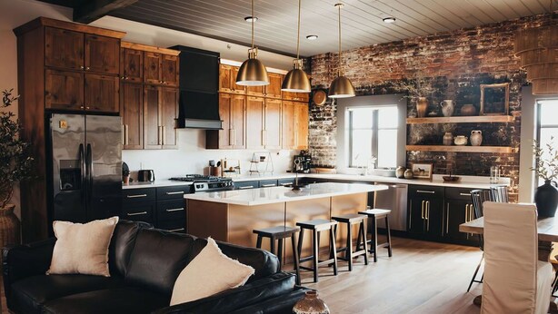 A modern kitchen with wooden cabinets, a central island, pendant lights, and a brick wall.