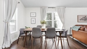 Un coin repas avec une table en bois et des chaises grises, un canapé en cuir marron et une fenêtre agrémentée de rideaux blancs.