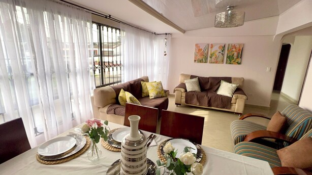 A living room with two sofas, a dining table set with plates and a vase of flowers, and a view through a window with curtains.