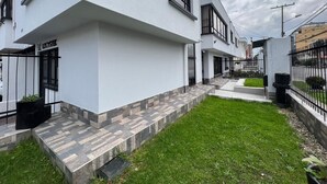 A modern two-story residential building with a flat roof, white exterior walls, and a small front yard with a paved pathway and a grassy area.