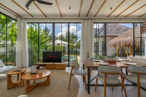 A modern living room with a large TV, comfortable seating, and a view of a tropical outdoor area. - Beta Villa by DH (Ubud)