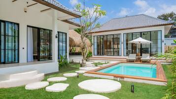 Una casa moderna con piscina, una construcción con techo de paja y un jardín cuidado con piedras a modo de camino.