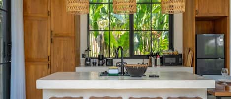 A modern kitchen with a large island, wooden bar stools, and a view of lush greenery through the window.