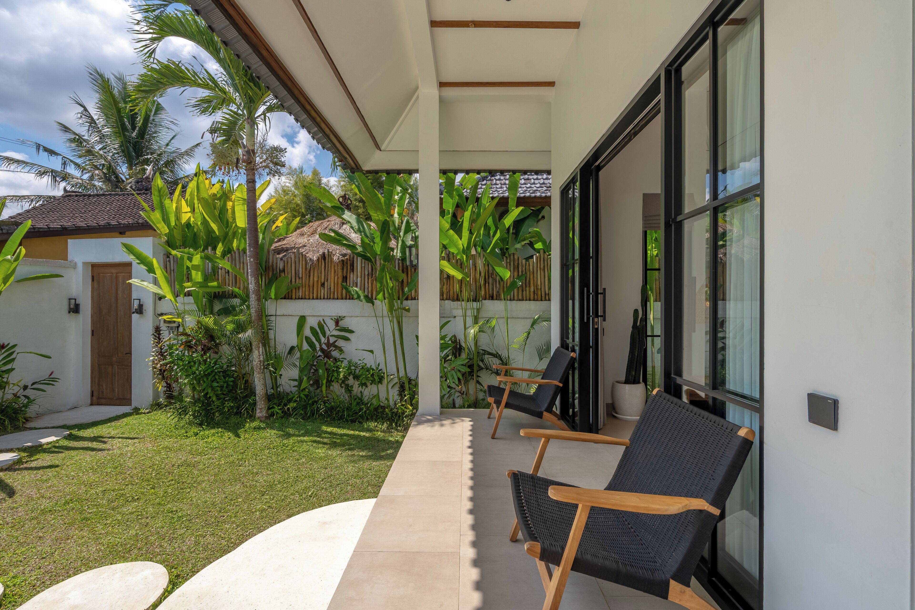 A covered outdoor seating area with two chairs, a concrete pathway, and lush greenery including banana plants.