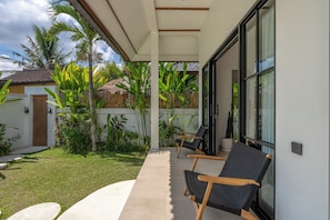 A covered outdoor seating area with two chairs, a concrete pathway, and lush greenery including banana plants. - Beta Villa by Betterplace (Ubud)