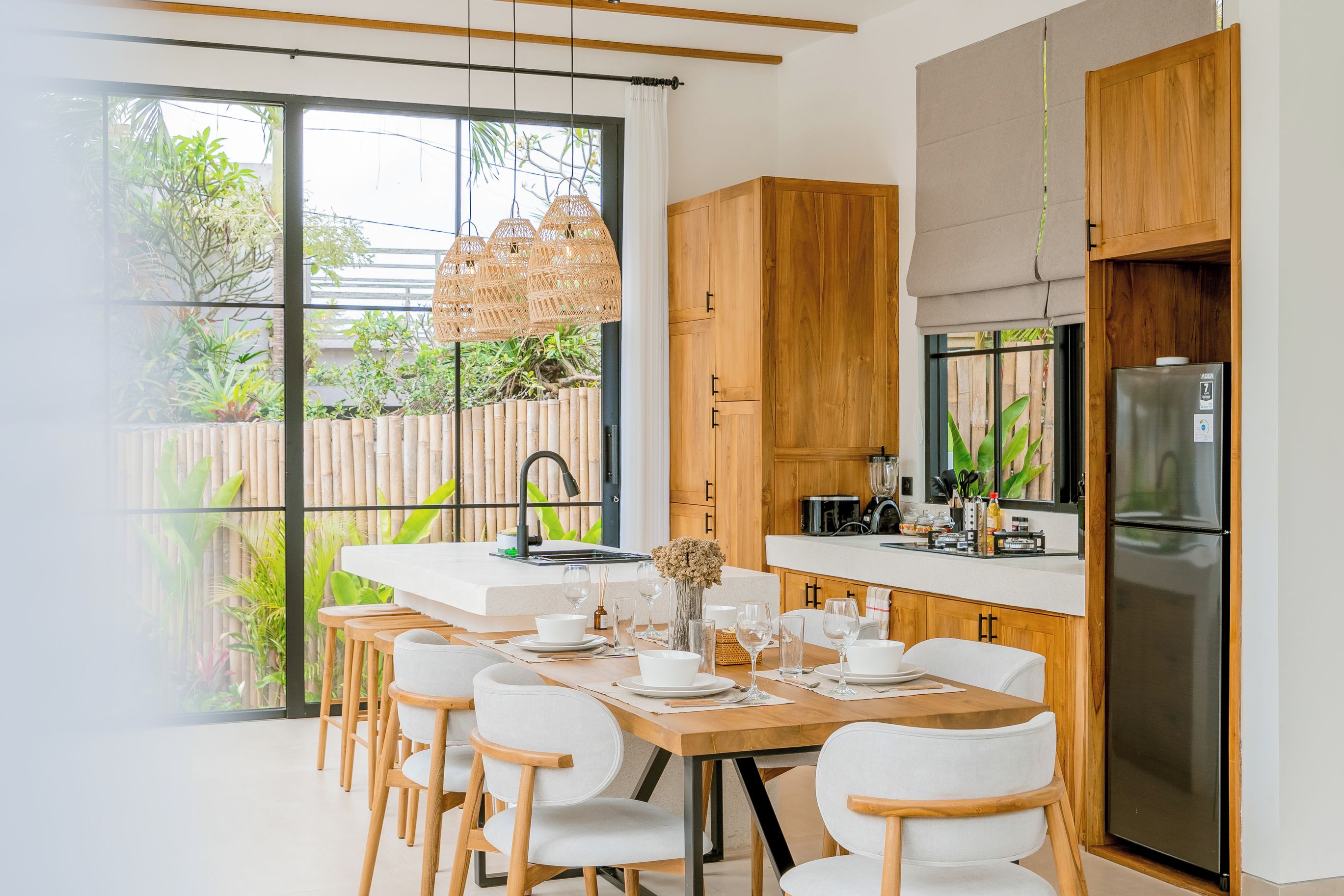 A modern kitchen with wooden cabinets, a stainless steel refrigerator, and a dining area with white chairs and a wooden table.