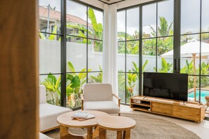 A modern living room with a large TV, comfortable seating, and a view of a swimming pool and lush greenery. - Beta Villa by DH (Ubud)