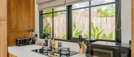 A modern kitchen with a built-in stove, a microwave, and a window with a view of a bamboo fence and greenery.