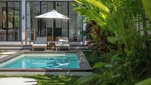 A swimming pool with lounge chairs and an umbrella, surrounded by lush greenery and a modern building with large windows.