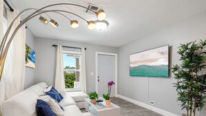 A modern living room with a white sofa, a coffee table with books and plants, a large window with curtains, and a painting of a beach scene.