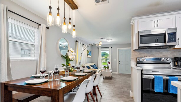 A modern kitchen with stainless steel appliances, a dining area with a wooden table and white chairs, and a large window with blinds.