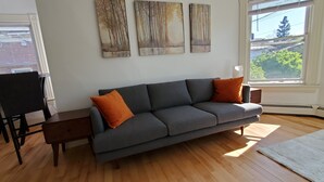 A living room with a gray sofa, two orange cushions, a wooden coffee table, and a white rug. There are two black chairs, a small wooden side table, and three abstract paintings on the wall. A window provides a view of trees and a building. - Stylish Studio in the Queen City
 (Burlington)