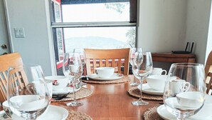 Una mesa de comedor dispuesta con platos, fuentes y copas de vino, con vista al exterior a través de la ventana.