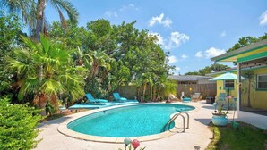 Un patio trasero con piscina, sillas de descanso y plantas tropicales.