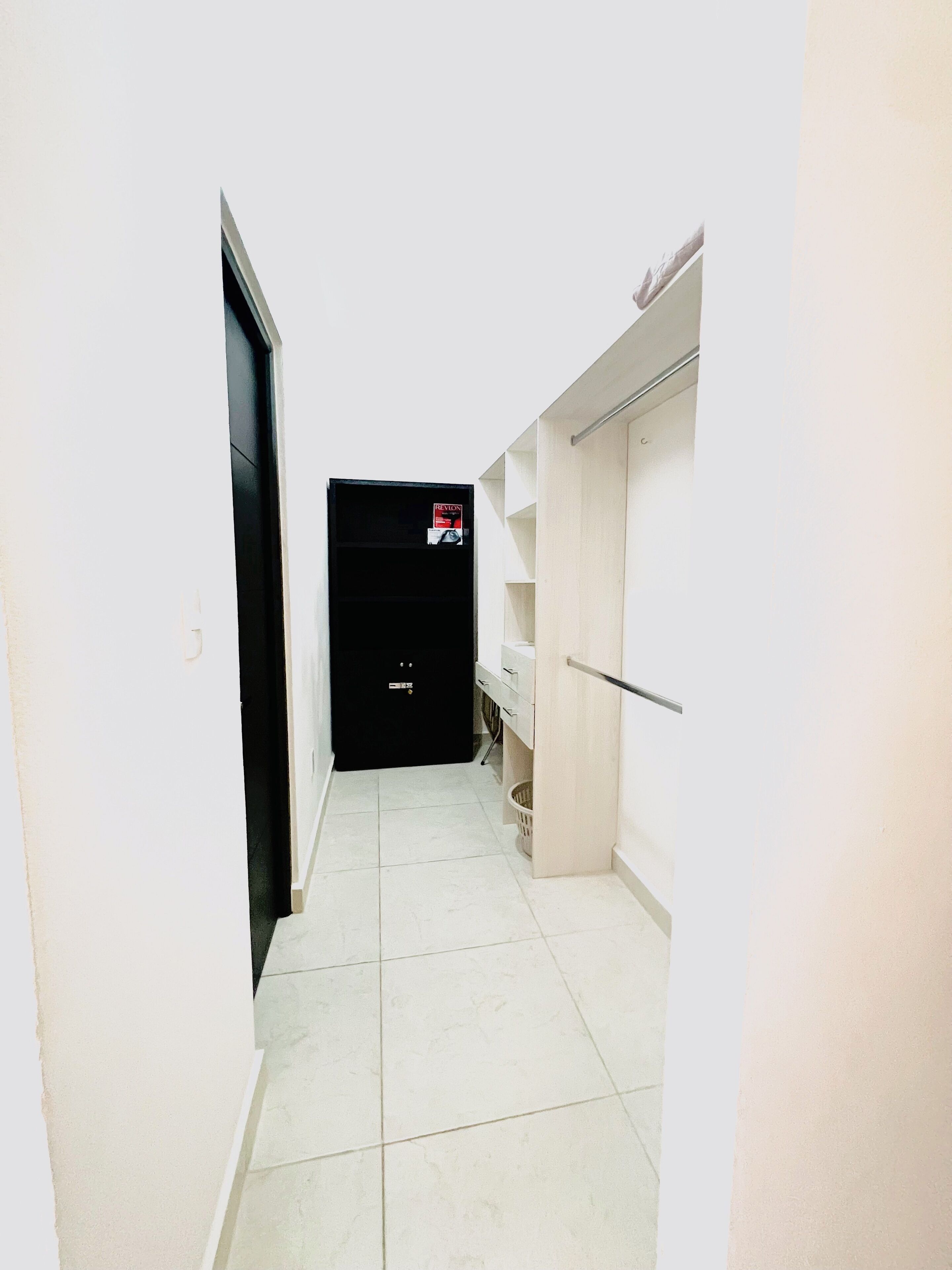 Un couloir étroit, avec une porte noire au fond, des murs blancs et un sol carrelé.