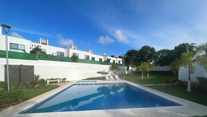 A rectangular swimming pool with a clear blue water surface, surrounded by a concrete deck. There are two white lounge chairs and a small umbrella near the pool. The pool is set against a backdrop of a white building with a green roof and a clear blue sky above. - Spacious Cancun Retreat • Pool • Sleeps 6 • Kid Friendly (cancun)
