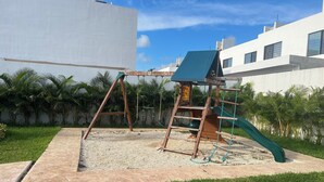 A playground with a swing set, slide, and climbing structure in a grassy area with a white building in the background. - Spacious Cancun Retreat • Pool • Sleeps 6 • Kid Friendly (cancun)