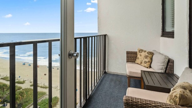 Un balcón con vista a la playa, mobiliario de mimbre y una ventana con persianas.