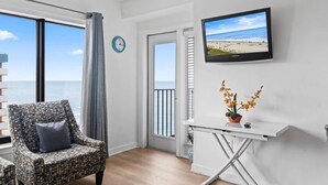 Una habitación con vista a la playa, una silla, una mesa con una planta y un televisor en la pared.
