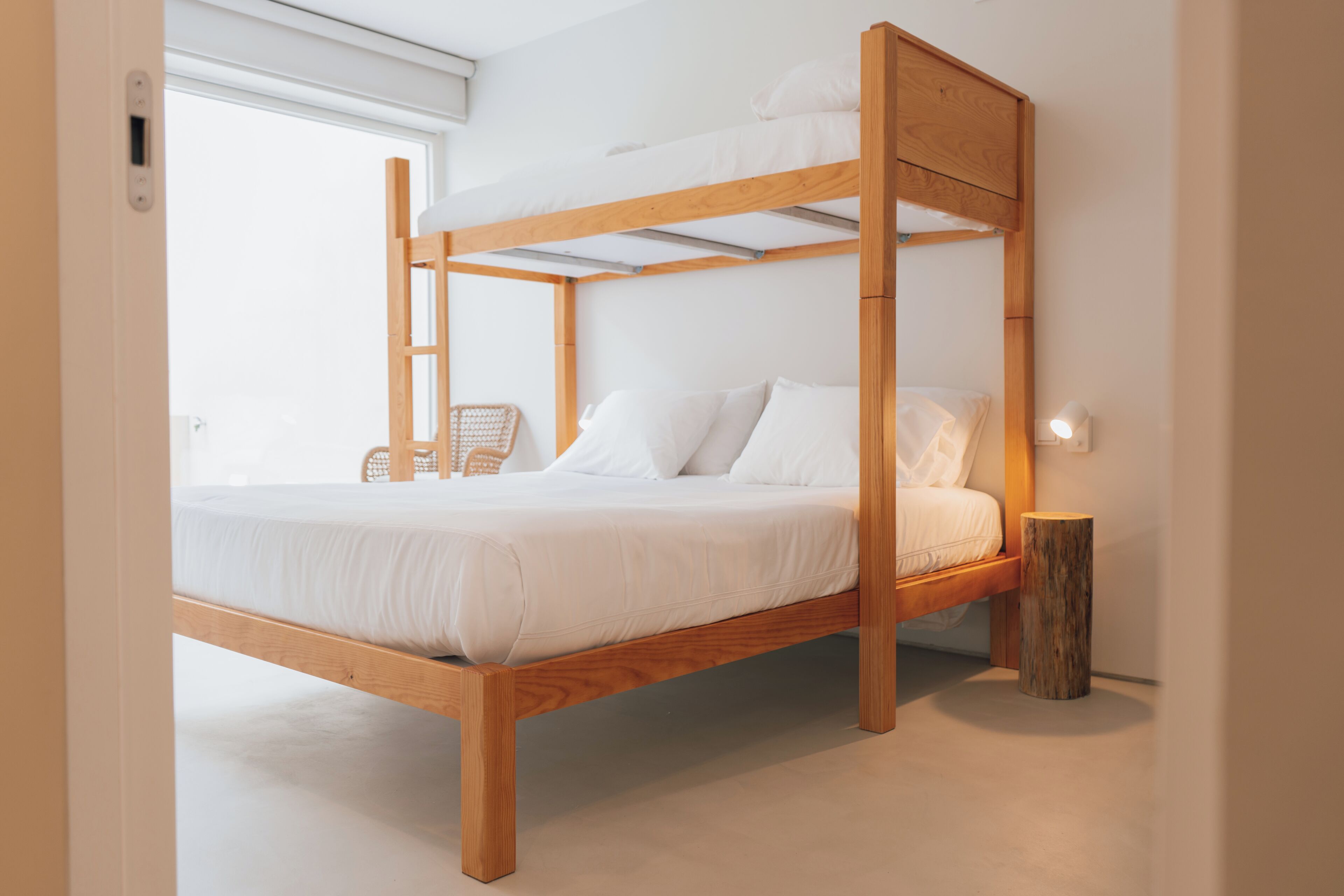 A wooden bunk bed with white bedding, a wooden nightstand, and a wicker chair.