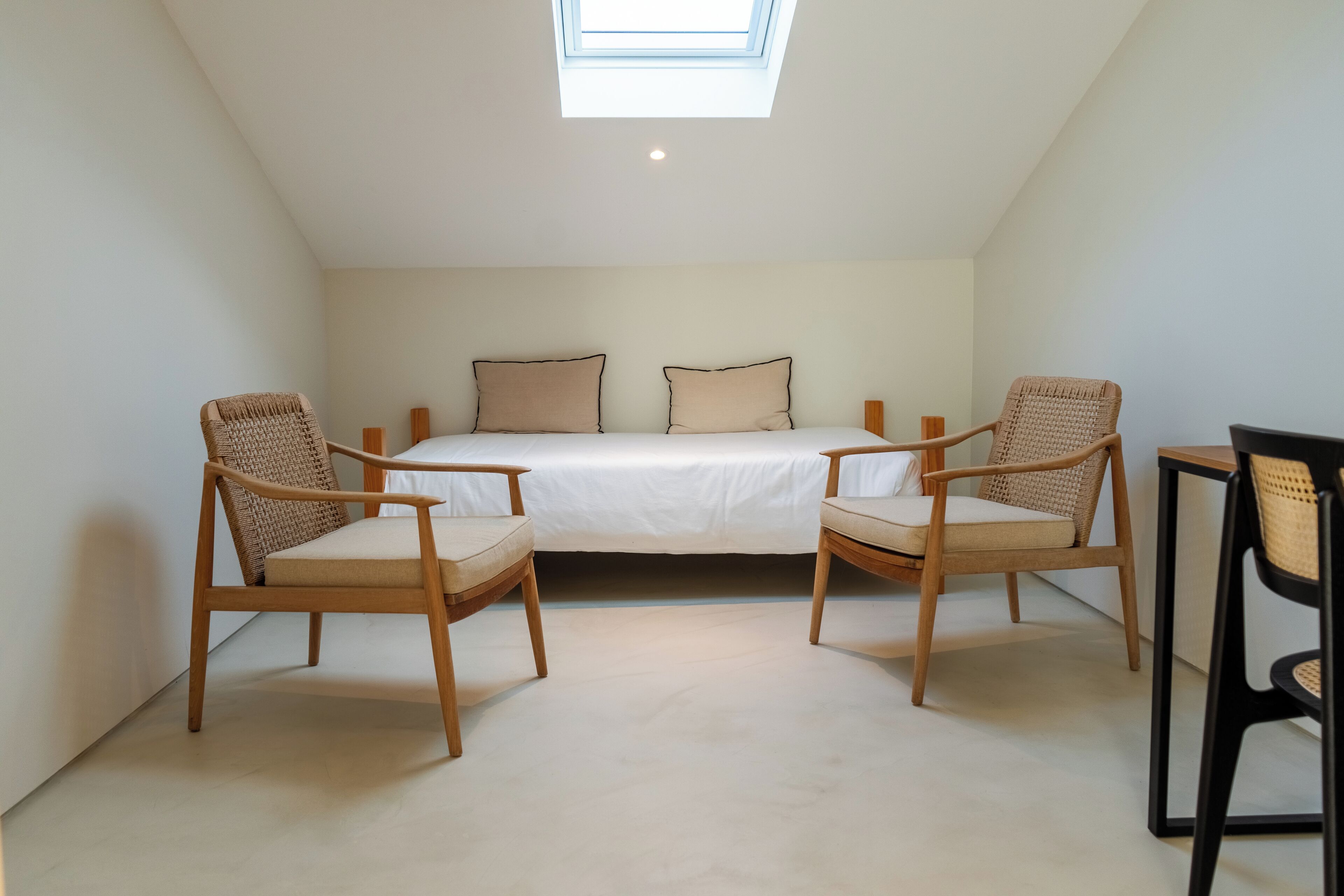 A bedroom with a skylight, two wooden chairs, a bed with white bedding, and a small table.