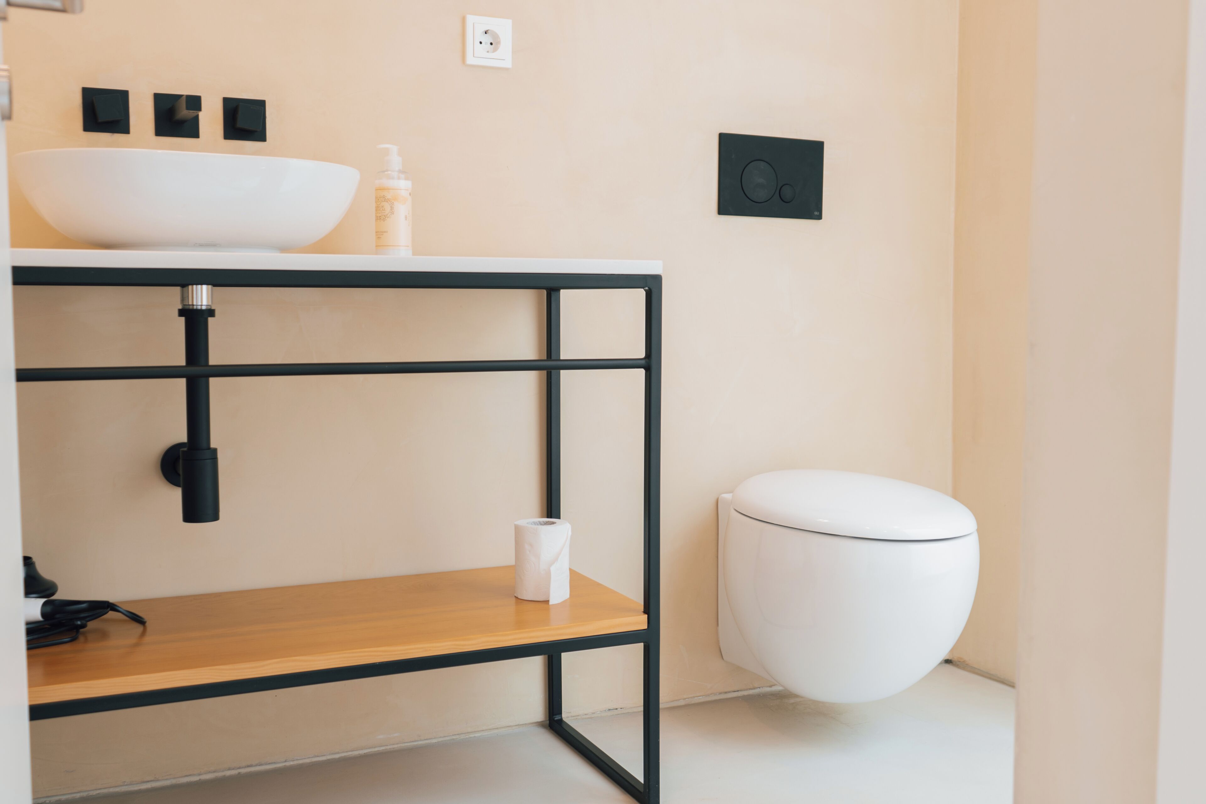 Une salle de bain moderne avec un lavabo blanc, un cadre en métal noir, une étagère en bois, des toilettes et un interrupteur mural.