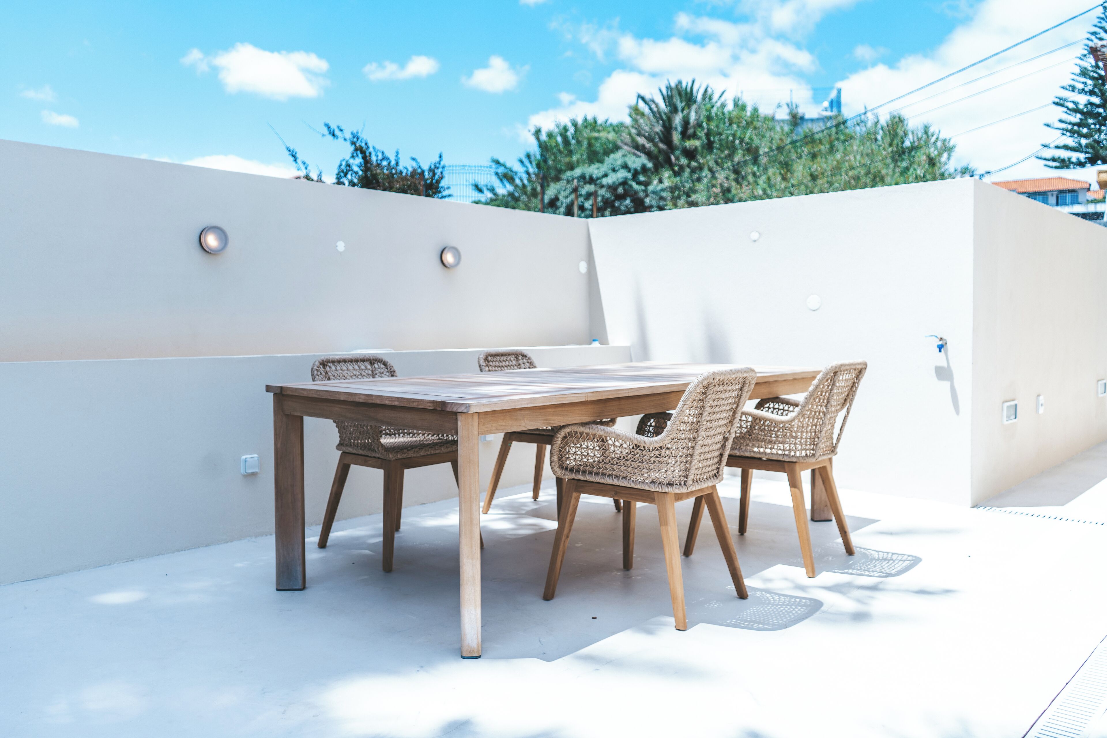 Un patio al aire libre con una mesa de madera y sillas de mimbre, rodeado por un muro blanco y un cielo despejado.