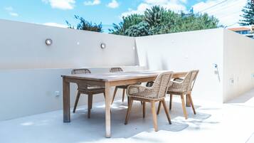 Un patio al aire libre con una mesa de madera y sillas de mimbre, rodeado por un muro blanco y un cielo despejado.