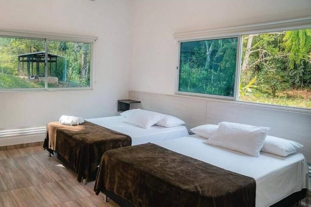A room with two beds, a standing fan, and large windows with a view of greenery.