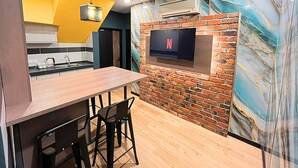 A modern kitchen with a wooden dining table, black chairs, a hanging light fixture, and a TV mounted on a brick wall. - Seeds Hotel Cheras Maluri (Kuala Lumpur)