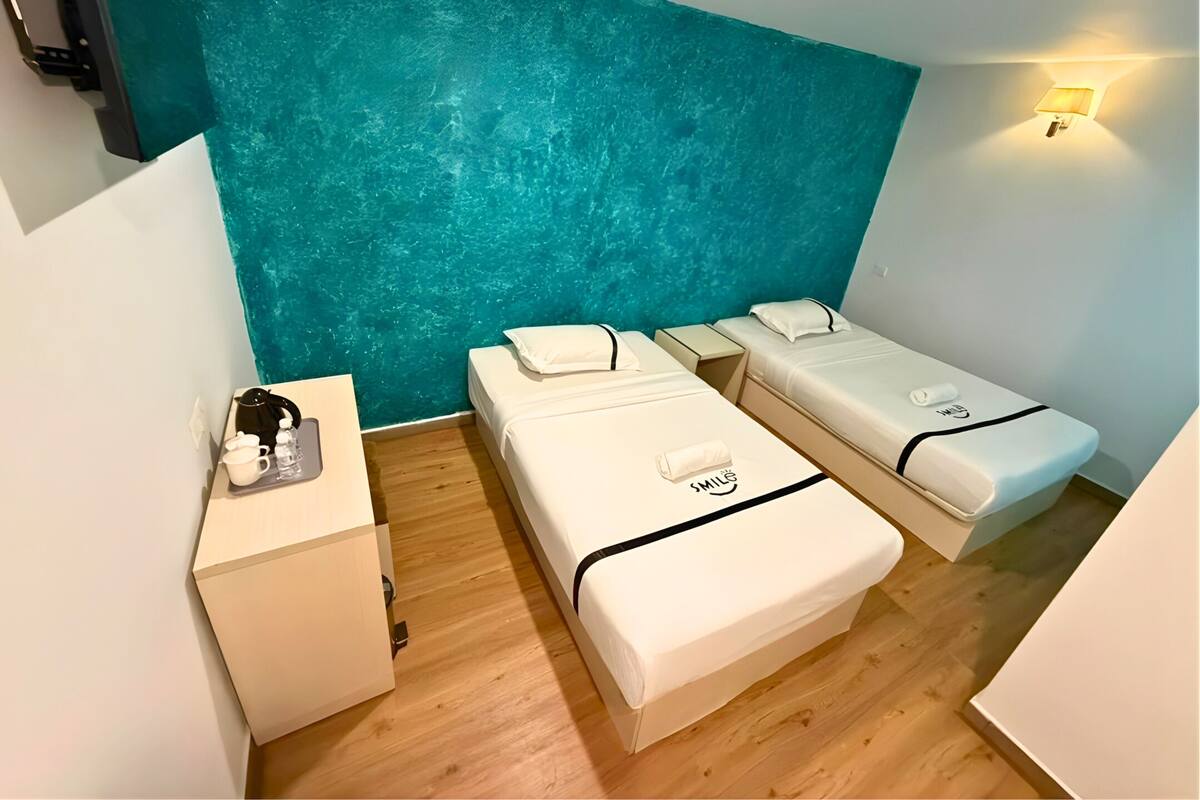 Une chambre d’hôtel avec deux lits simples, une petite table avec une bouilloire et des tasses, et un mur d’accent de couleur turquoise.