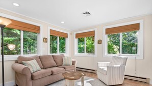 Un salon spacieux avec un canapé beige, un fauteuil à rayures et une table basse. De grandes fenêtres équipées de stores en bambou offrent une vue sur la verdure environnante.
