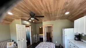 Una habitación con techo de madera, una cama, un refrigerador blanco, una cafetera y un sofá con un cojín estampado.