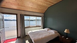 A bedroom with a wooden ceiling, a bed with white bedding, a nightstand with a lamp, and a view of a lake through a large window.